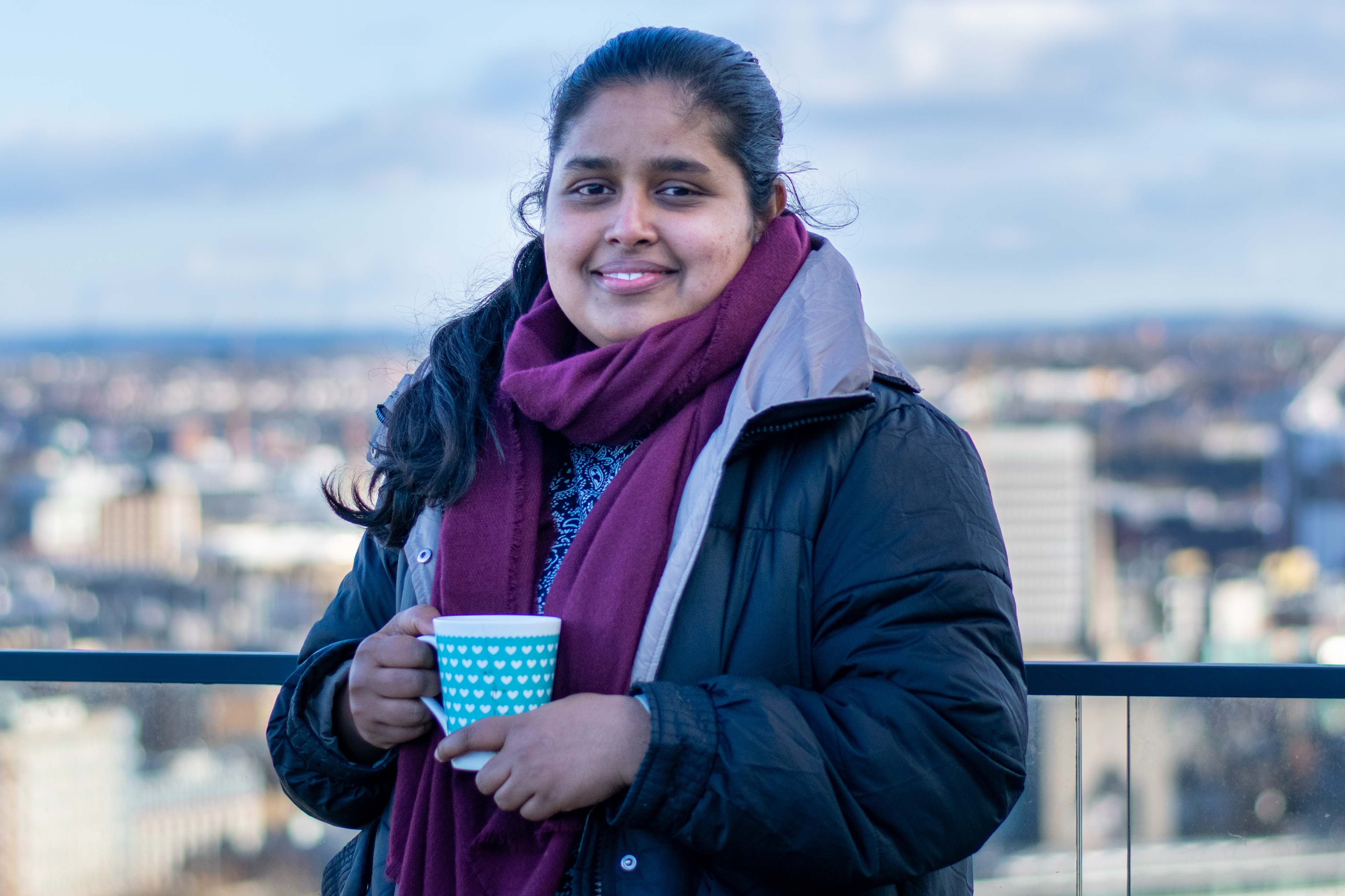 An Asian lady stands in Bristol with a purple scarf and blue coat holding a cup of tea